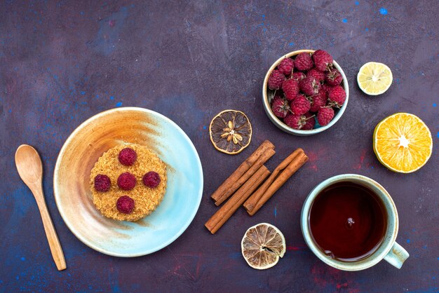 Top view of little round cake with fresh raspberries inside plate with fruits cinnamon on the dark surface