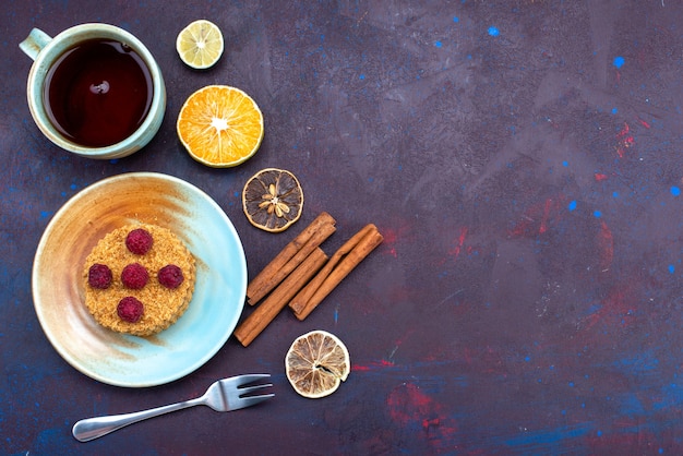 Top view of little round cake with fresh raspberries inside plate with tea on the dark-blue surface