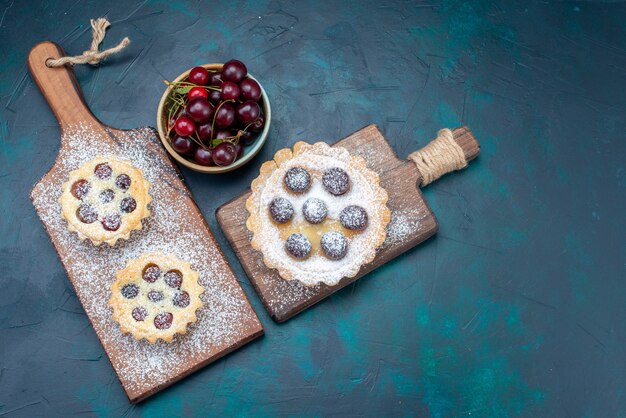 Top view little tasty cakes with sugar powder fruits along with fresh sour cherries on the dark background cake biscuit sugar sweet bake