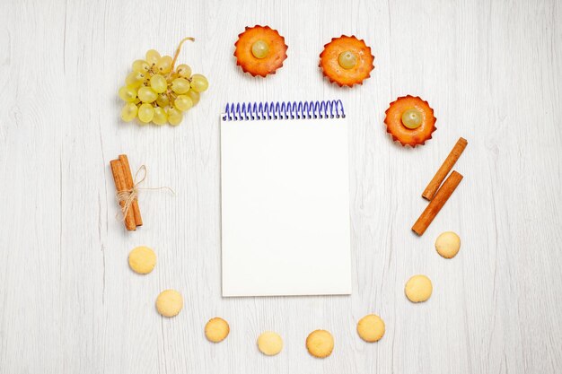 Top view little yummy cakes with grapes notepad and cookies on white desk fruit cake biscuit sweet dessert tea