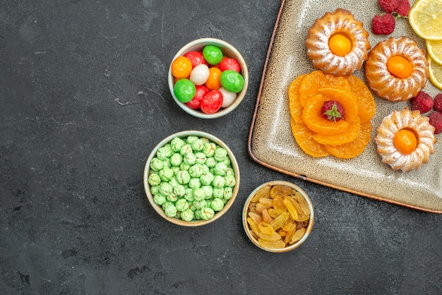 Top view of little yummy cakes with lemon slices tangerines and candies on dark