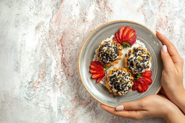 Вид сверху маленькие вкусные пирожные с клубникой на белой поверхности, празднование дня рождения, сладкий бисквитный торт