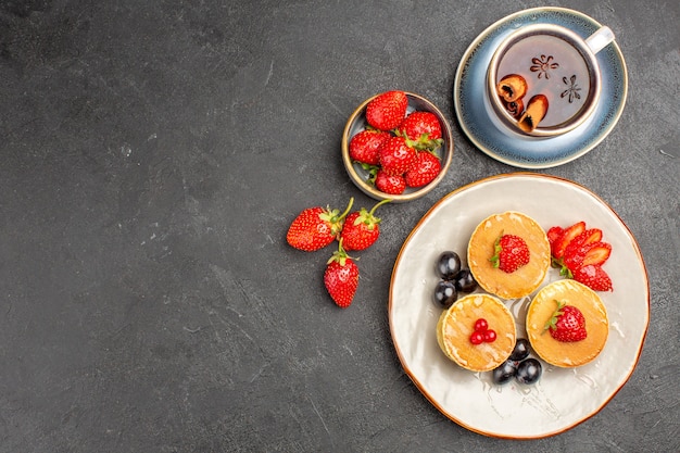 Foto gratuita vista dall'alto piccole frittelle gustose con frutta e tazza di tè sulla superficie grigia torta torta di frutta