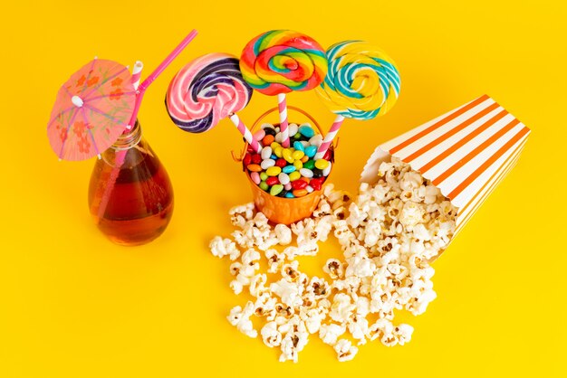 Вид сверху леденцы на палочке и попкорн с коктейлем и разноцветными конфетами на желтом фоне пьют сахарный конфитюр