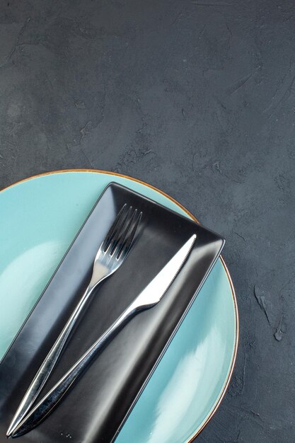 Top view long black plate with blue plate fork and knife on dark surface
