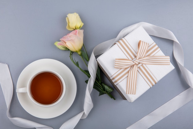 Top view of lovely flowers with white gift box ribbon and a cup of tea on a gray background