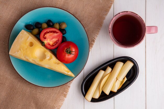 Top view maasdam cheese with tomatoes and olives on a blue plate with a cup of tea and smoked cheese on a white plate