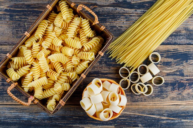 Top view macaroni in basket and bucket with pasta and spaghetti on dark wooden background. horizontal