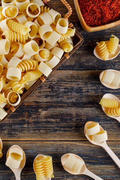 Top view macaroni pasta in basket and spoons with red spice on wooden background. vertical