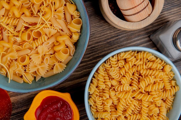 Top view of macaronis as rotini and others in bowls with ketchup salt black pepper on wood