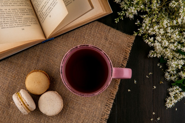 Vista dall'alto di macarons con una tazza di tè su un tovagliolo beige con un libro aperto e fiori