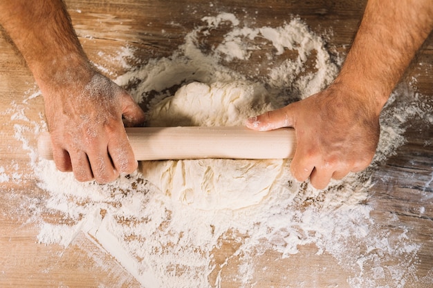 木製のテーブルにローリングピンで生地を平らにする男性のパン屋の手のトップビュー