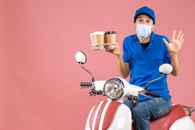 Top view of male delivery person in mask wearing hat sitting on scooter delivering orders feeling shocked on peach background