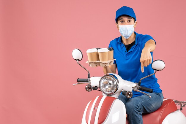 Free photo top view of male delivery person in mask wearing hat sitting on scooter delivering orders pointing down on peach background