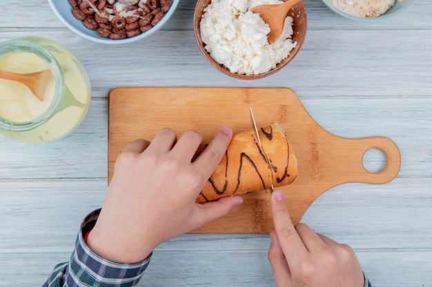 Top view of male hands cutting roll with knife on cutting board with condensed milk cottage cheese cereals on wooden board