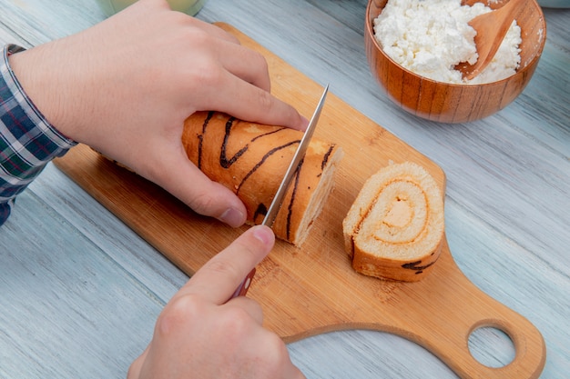 Top view of male hands cutting roll with knife on cutting board with cottage cheese on wooden board