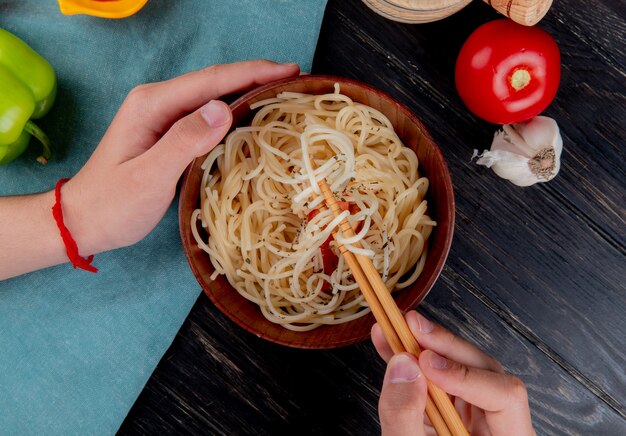 Top view of male hands holding chopsticks and bowl with macaroni pasta with tomato pepper garlic on wooden surface