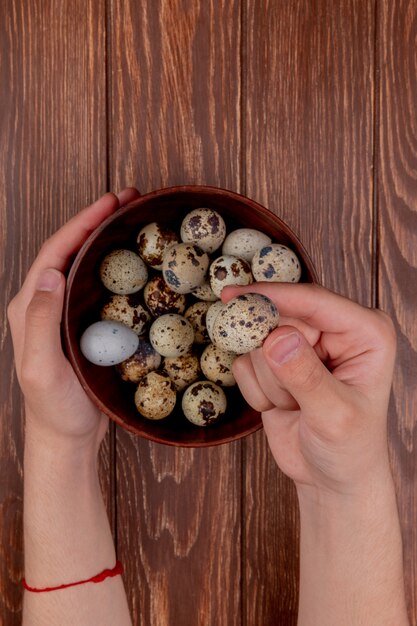 Вид сверху на мужские руки, держащие перепелиные яйца на деревянной миске с несколькими перепелиными яйцами на деревянном фоне