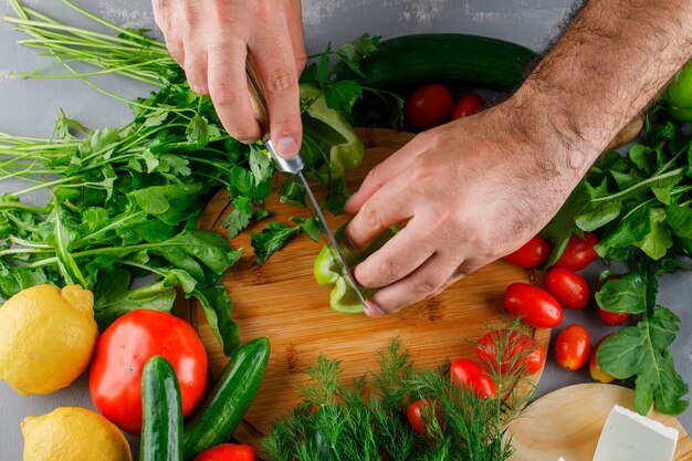 Top view man slicing green pepper on cutting board with tomatoes, salt, cheese, lemon on gray surface