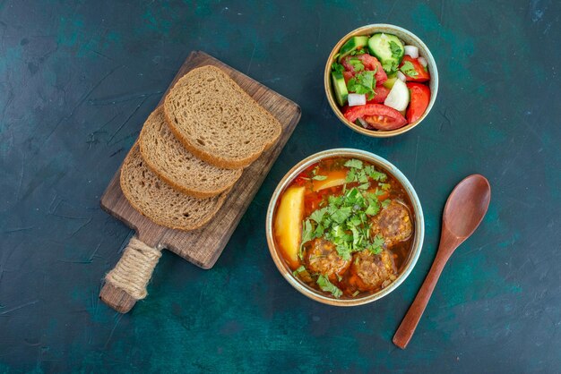 Top view meat soup with meatballs greens and sliced potatoes with bread loafs on the dark blue desk