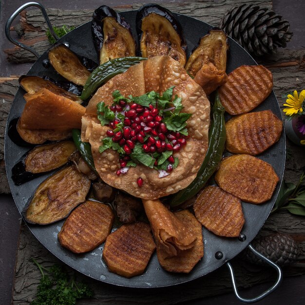 Вид сверху мясо с печеным картофелем, баклажанами, помидорами, перцем и украшенное гранатом на деревянной коре