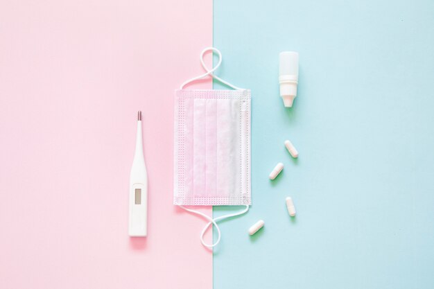 Top view of medical mask, pills and thermometer on pink and green background.