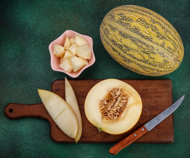 Top view of melon slices on a wooden kitchen board with peels with knife on green surface