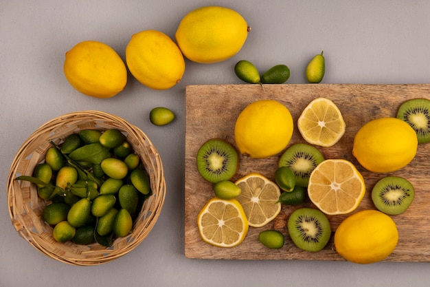 Вид сверху слегка сладких кинканов на ведре с лимонами и киви, изолированными на деревянной кухонной доске на серой стене