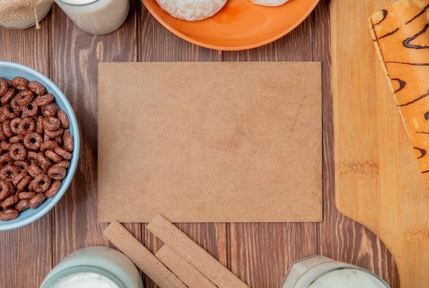 Top view of milk products as milk cream sour clotted milk yogurt soup with cereals cookies gingerbreads and roll on cutting board around cardboard on wooden background with copy space