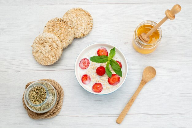 Top view milk with oatmeal inside plate with strawberries along with crackers and honey on white, breakfast cereals health