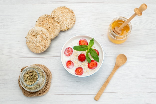 Top view milk with oatmeal inside plate with strawberries along with crackers and honey on white, breakfast cereals health