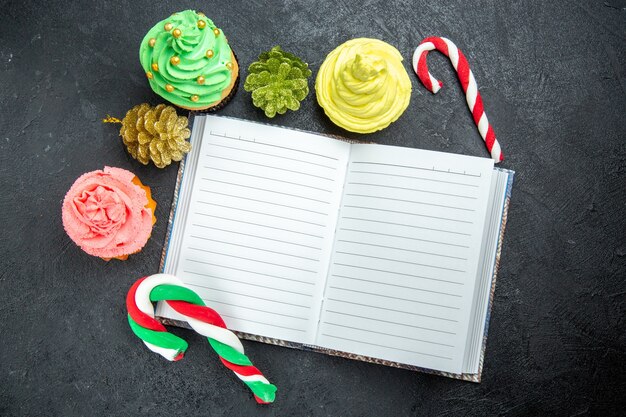 Top view mini colorful cupcakes a notebook xmas candies and ornaments on dark surface