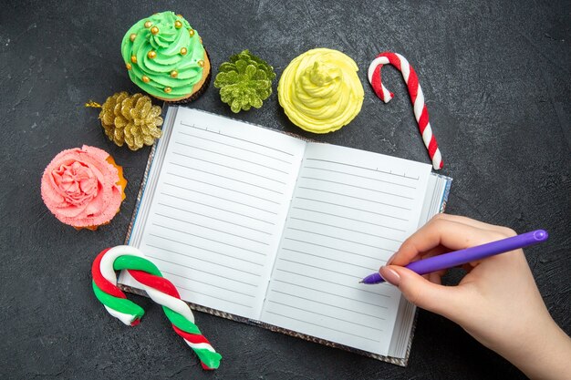 상위 뷰 미니 다채로운 컵케이크 노트북 크리스마스 사탕과 장식품 펜은 어두운 표면에 있는 여성의 손에 있습니다.