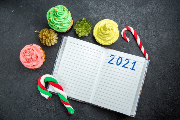 Top view mini colorful cupcakes written on notebook xmas candies and ornaments on dark background