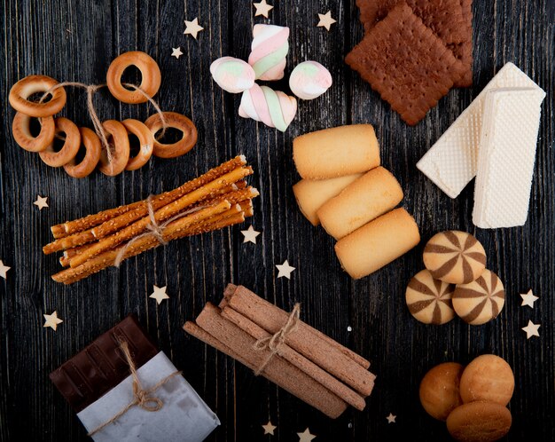 Top view mix of cookies with muffins waffles marshmallows and bread sticks on a black wooden background