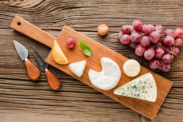 Top view mix of gourmet cheese on wooden cutting board with grapes and ustensils