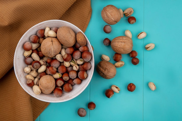 Top view of mix nuts in a bowl with walnuts hazelnuts with pistachios with a brown towel on a turquoise surface