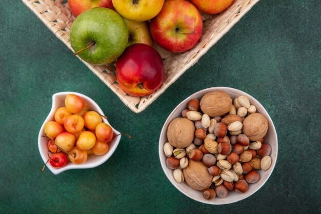 Top view of mix nuts with white cherries and apples in a basket on a green surface