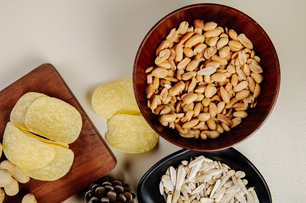 Вид сверху смеси соленых закусок к пиву арахис в деревянной миске картофельные чипсы и семечки на белом