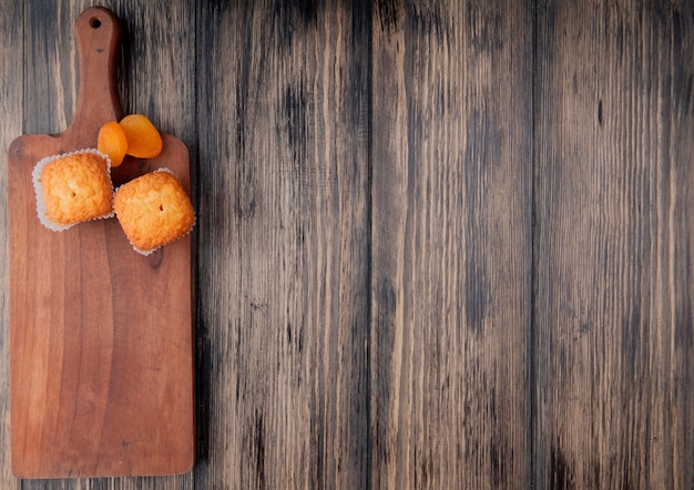 top view of muffins and dried apricots on wooden cutting board on rustic surface with copy space