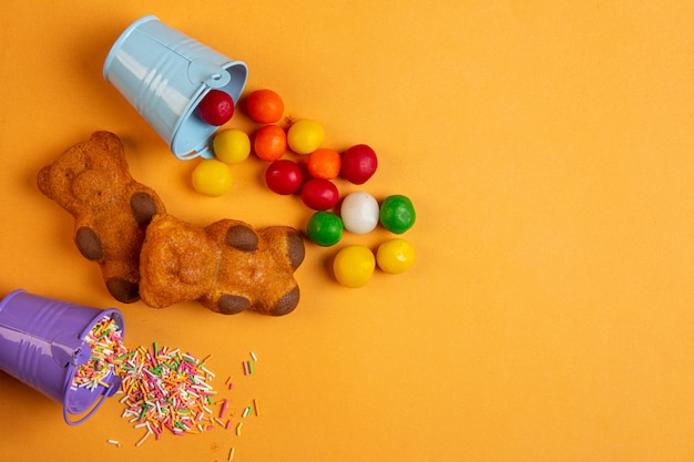 Top view of multicolor chocolate candies scattered from small bucket and sponge cakes in a shape of bear on yellow