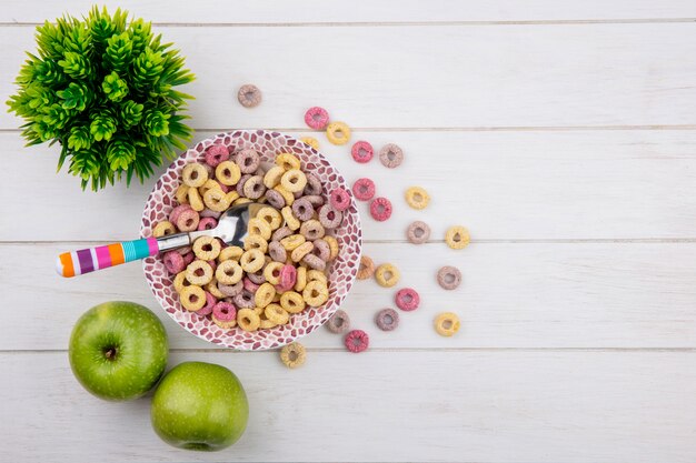 Top view of multicolored cereals on a bowl with a spoon with green apples on white surface
