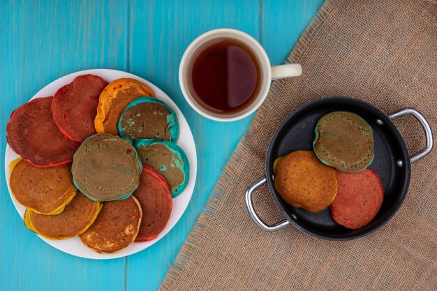 Top view multicolored pancakes on a plate and in a frying pan with a cup of tea on a beige napkin on a turquoise background