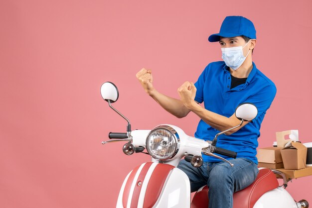 Vista dall'alto dell'uomo corriere nervoso in maschera medica che indossa un cappello seduto su uno scooter su sfondo color pesca pastello