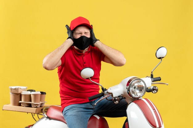 Top view of nervous young adult wearing red blouse and hat gloves in medical mask delivering order sitting on scooter feeling emotional