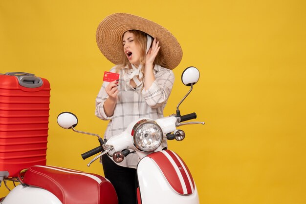 Top view of nervous young woman wearing hat collecting her luggage sitting on motorcycle and holding bank card