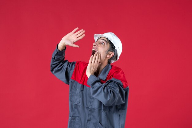 Top view of nervous young worker in uniform with hard hat and looking up on isolated red wall