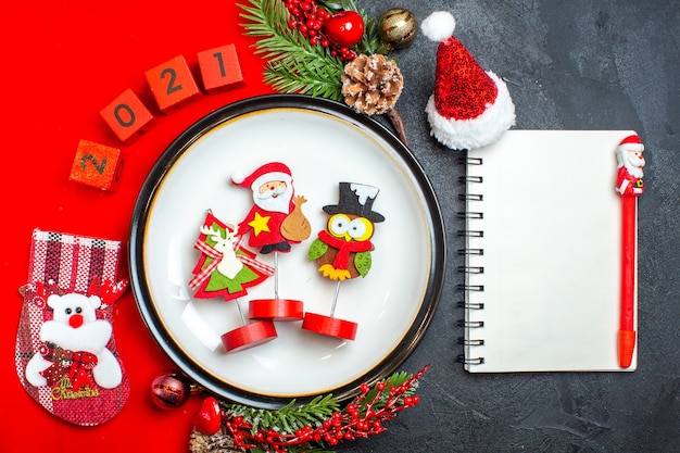 Free photo top view of new year background with dinner plate decoration accessories fir branches and numbers christmas sock on a red napkin notebook with pen on a black table