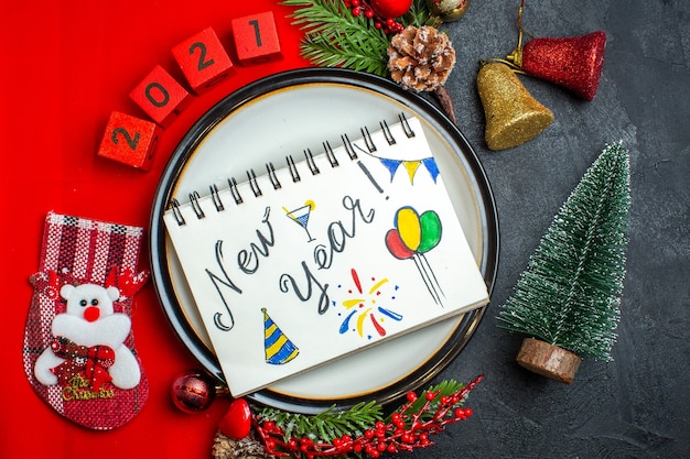 Top view of New year background with dinner plate decoration accessories fir branches and numbers on a red napkin next to christmas tree on a black table