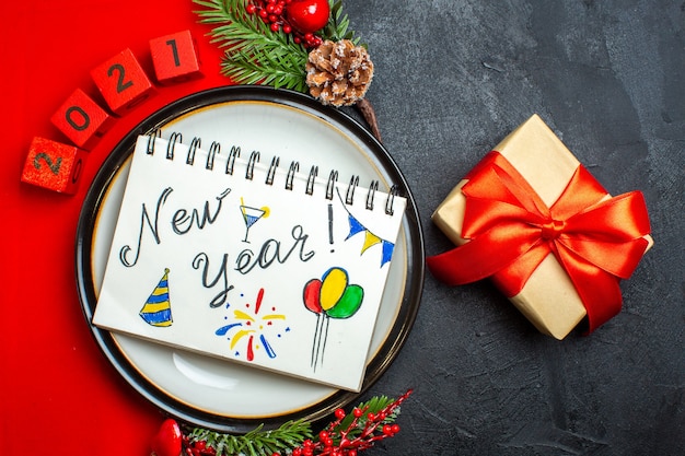 Вид сверху новогоднего фона с блокнотом с новогодними рисунками на обеденной тарелке, украшениями, еловыми ветками и числами на красной салфетке, и подарком на черном столе
